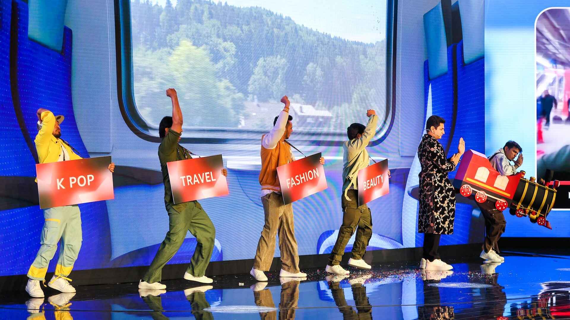 Performers on stage, joyfully lifting their arms, carrying red-coloured signs that say 'K-pop', 'Travel', 'Fashion' and 'Beauty', creating an energetic atmosphere.